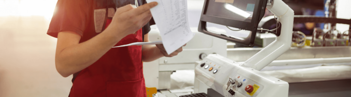 manufacturing employee standing at machine and looking at paperwork