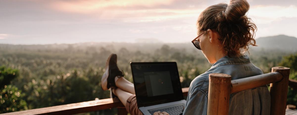 Woman remote working on laptop outside, with feet up on railing. 